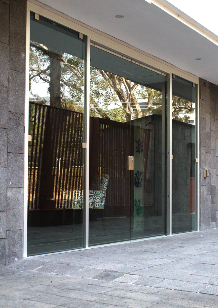 Puerta automática corrediza en vidrio a la entrada de un condominio vertical, tiene dos hojas móviles de apertura lateral y dos hojas fijas, todas las hojas en vidrio.