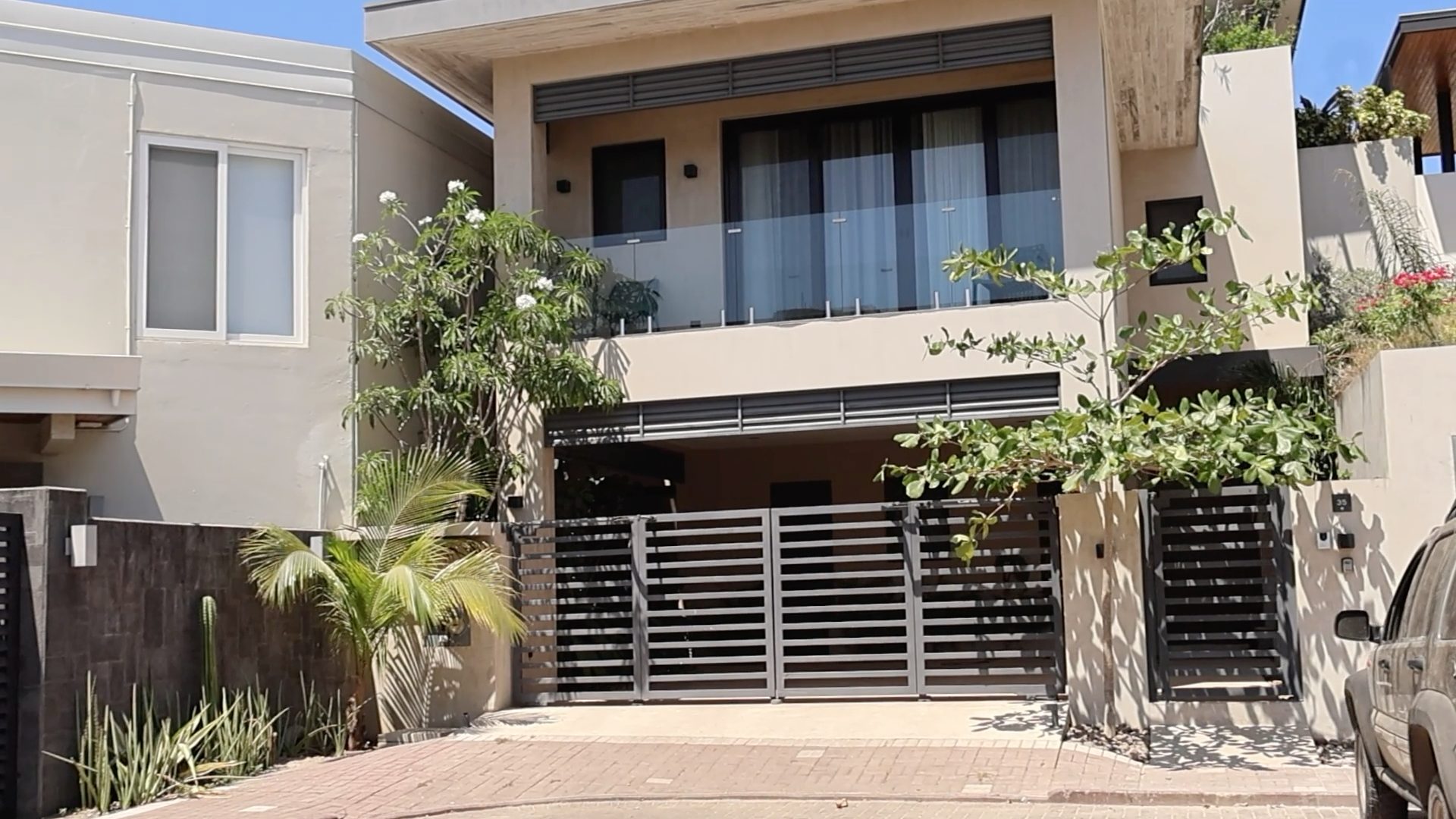 Vista frontal de la entrada a una casa de dos pisos, con vegetación y como eje central un portón abatible de dos hojas.